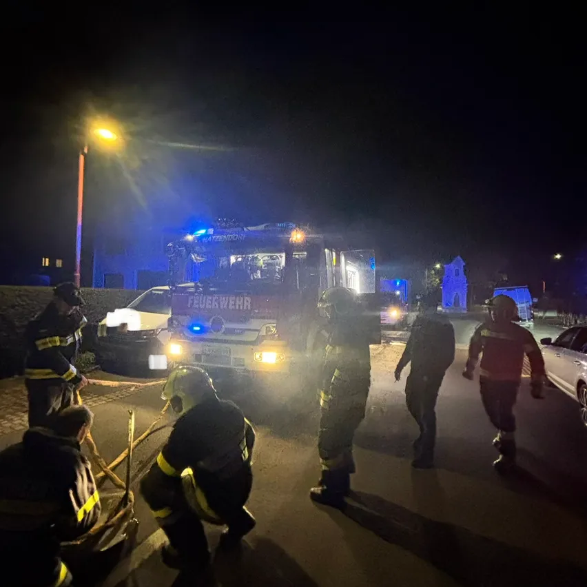 Eine Gruppe von Feuerwehrleuten reagiert auf einen Notfall bei Nacht. Sie tragen Schutzkleidung und Helme, und einer kniet auf dem Boden. Ein Feuerwehrwagen mit blinkenden Lichtern ist geparkt, und ein Auto ist am Straßenrand geparkt.