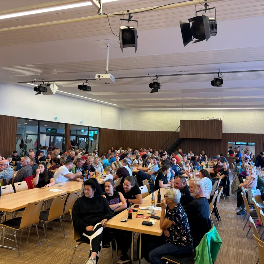 Eine große Gruppe von Menschen sitzt in einem Saal, einige stehen. Der Saal hat Holzböden, braune Wände und Deckenbeleuchtung.
