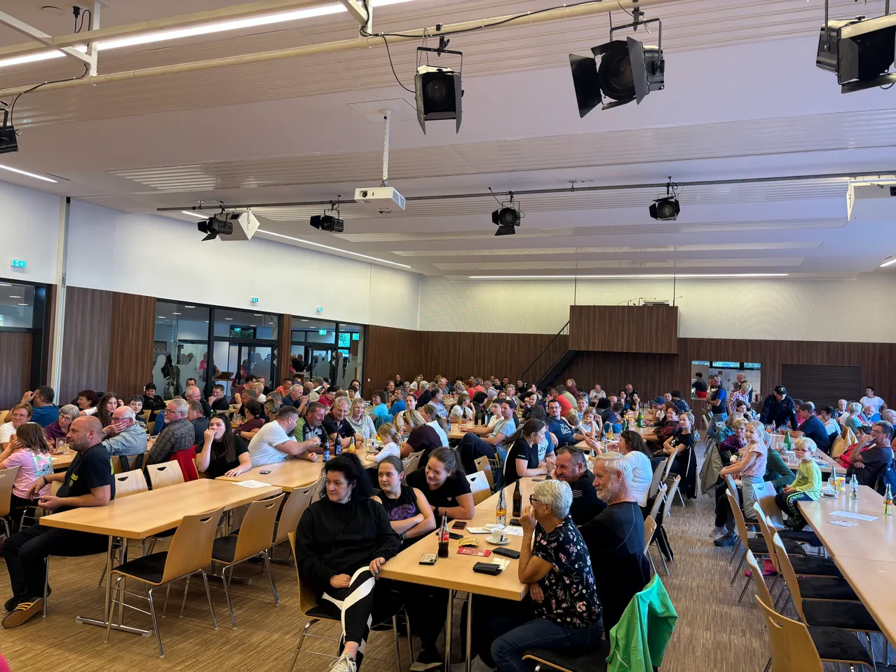 Eine große Gruppe von Menschen sitzt in einem Saal, einige stehen. Der Saal hat Holzböden, braune Wände und Deckenbeleuchtung.