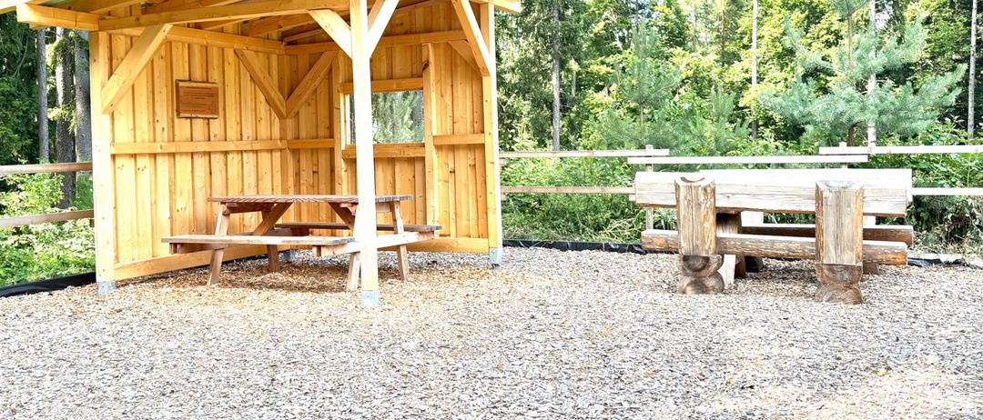 Ein hölzerner Pavillon mit Dach steht auf Kies, umgeben von Bäumen. Ein Picknicktisch mit Bänken befindet sich im Inneren. Der Bereich ist mit einer Holzbarriere eingezäunt.