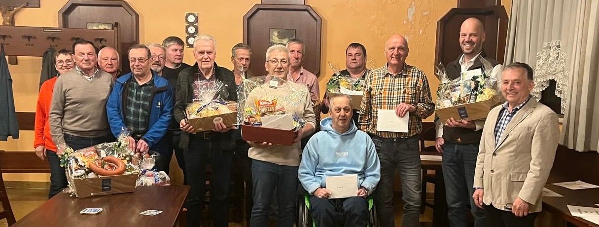 Eine Gruppe von Männern in einem Raum, einige halten Geschenke und lächeln. Ein Mann im Rollstuhl hält ein Papier. Dahinter hält ein Mann in einem blauen Mantel eine Geschenkbox. Eine Uhr und ein Bilderrahmen hängen an der Wand.