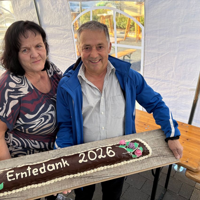Ein Mann und eine Frau stehen nebeneinander und halten einen Kuchen mit der Aufschrift 'Erntedank 2026'.
