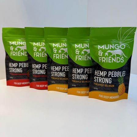 Fünf Packungen Mungo & Friends Hanf-Kieselsteine Stark sind auf einer weißen Oberfläche aufgereiht. Die Geschmacksrichtungen sind Erdbeer-Explosion, Ananas-Delirium und tiefe Erinnerungen.