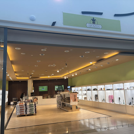 Ein offener Ladeneingang, der in einen Laden mit beleuchteter Decke und Vitrinen mit Produkten führt. Das Schild über dem Eingang heißt Grunzeug.