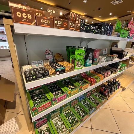 Ein Ladenregal mit einer Vielzahl von Produkten, darunter Kartons von O.C.B Slim, Hybrid und anderen Marken sowie Schachteln mit Cannabis-Lollies.