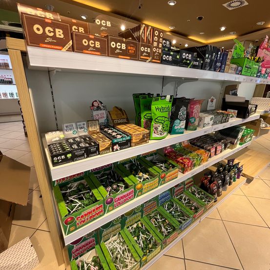 Ein Ladenregal mit einer Vielzahl von Produkten, darunter Kartons von O.C.B Slim, Hybrid und anderen Marken sowie Schachteln mit Cannabis-Lollies.