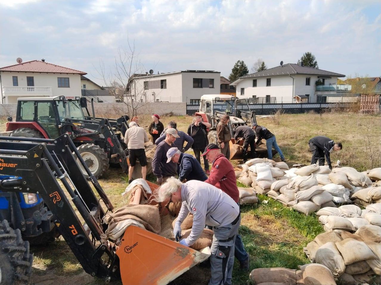 Eine Gruppe von Menschen lädt Sand in einen Bagger auf einem Feld mit Häusern im Hintergrund.