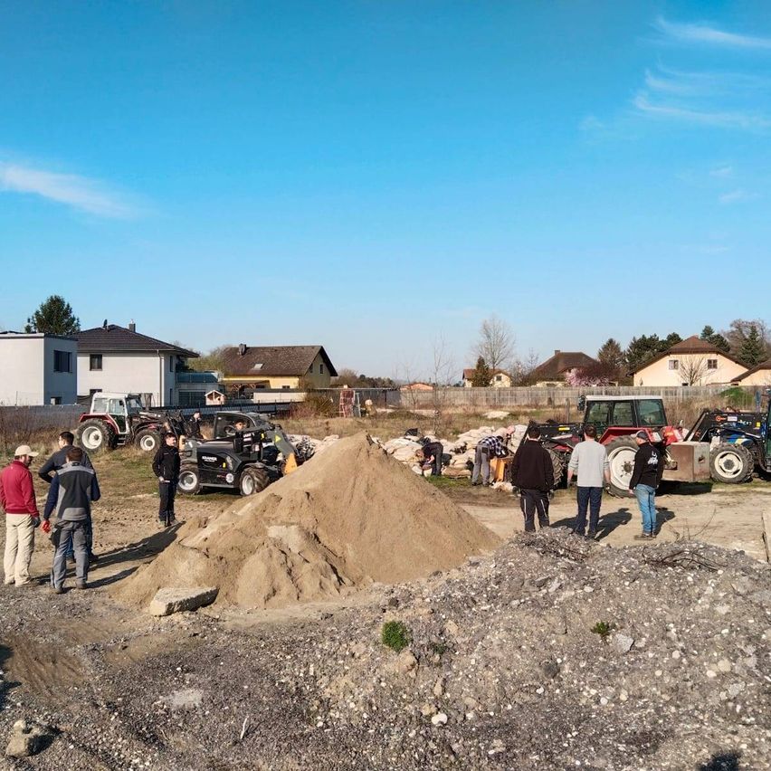 Eine Gruppe von Menschen versammelt sich um einen Haufen Erde auf einer Baustelle, mit einigen Traktoren und Maschinenfahrzeugen im Hintergrund.