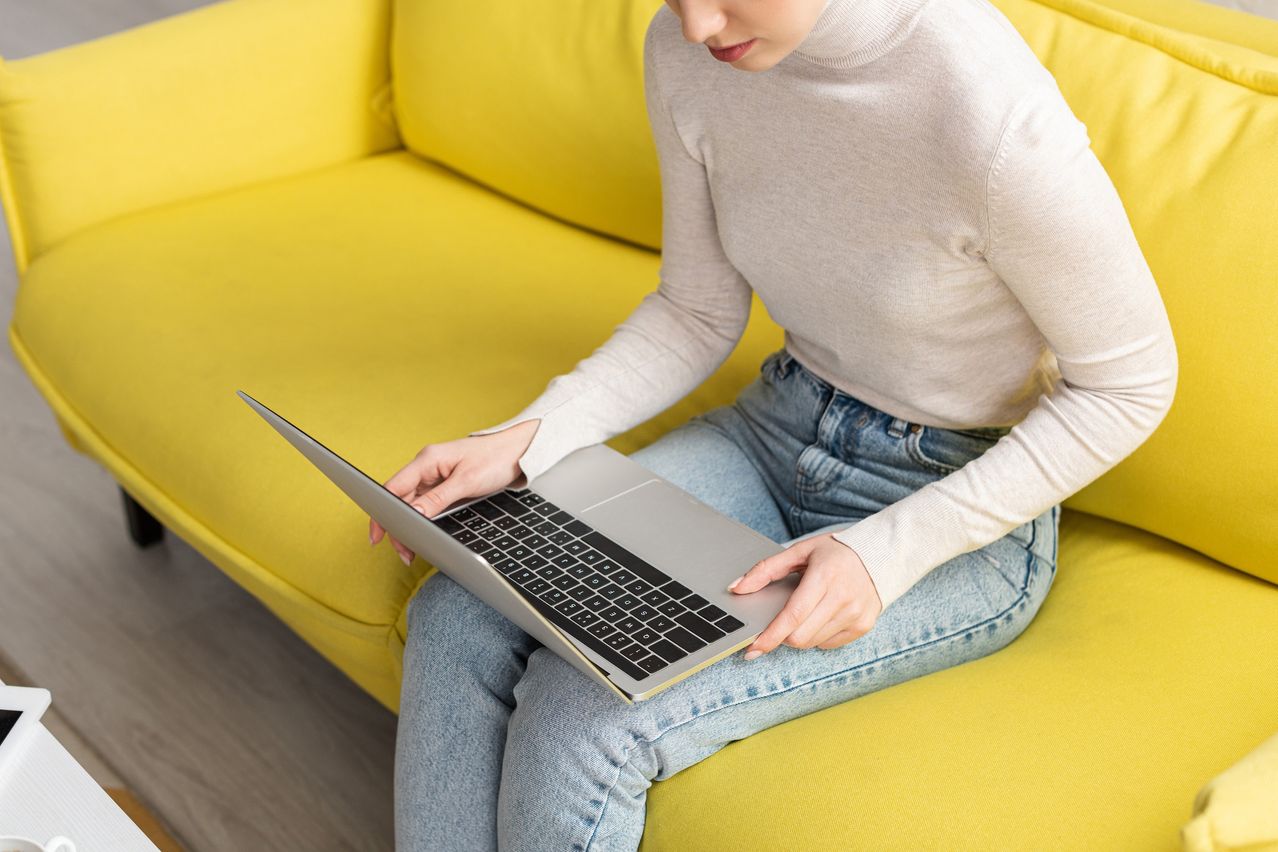 Eine Frau sitzt auf einem gelben Sofa und hält einen Laptop auf dem Schoß, der Bildschirm ist offen und ihre Finger sind auf der Tastatur.