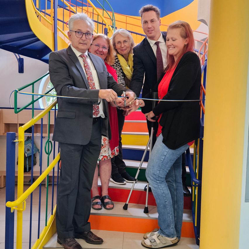 Eine Gruppe formell gekleideter Menschen steht auf einer Treppe und schneidet ein Band durch, wahrscheinlich während einer Eröffnungszeremonie.