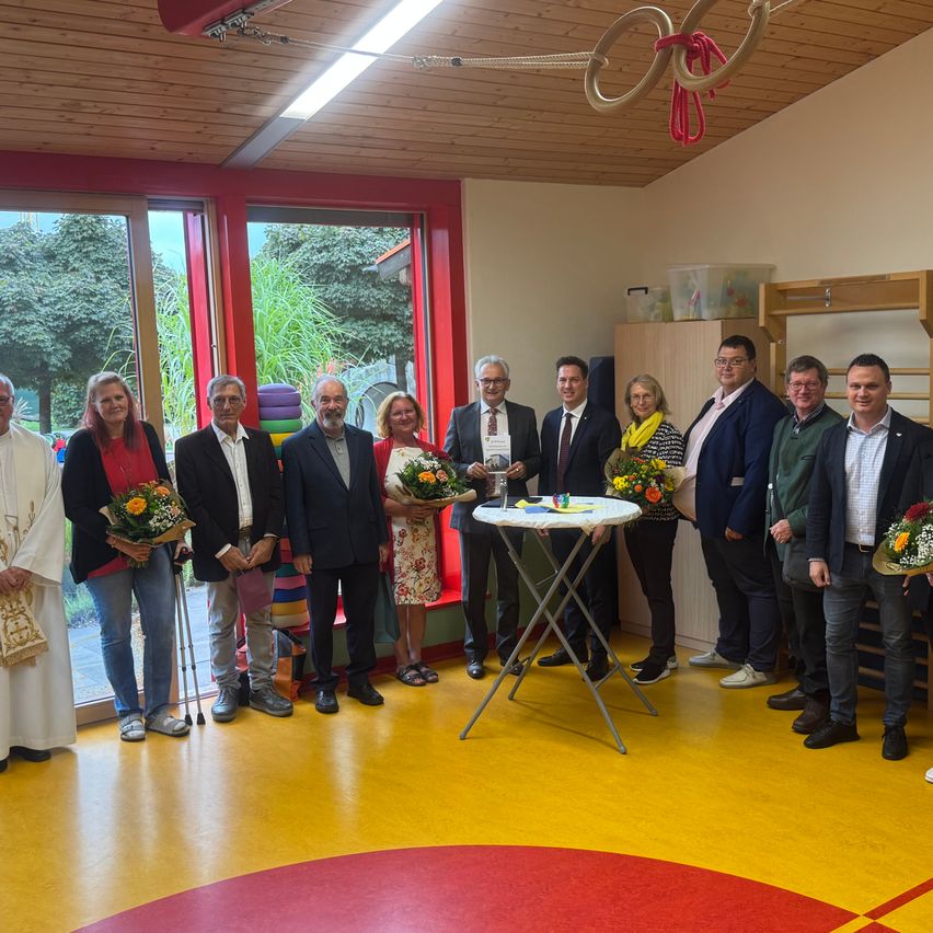 Eine Gruppe von Menschen steht in einem Raum mit einem Tisch, der Blumen und Urkunden hält, einige halten Blumensträuße. Ein Priester und mehrere Männer und Frauen in formeller Kleidung sind anwesend.