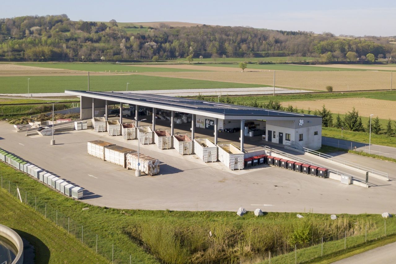 Luftaufnahme einer modernen Abfallanlage mit Solarpanelen auf dem Dach, umgeben von grünen Feldern und Hügeln.