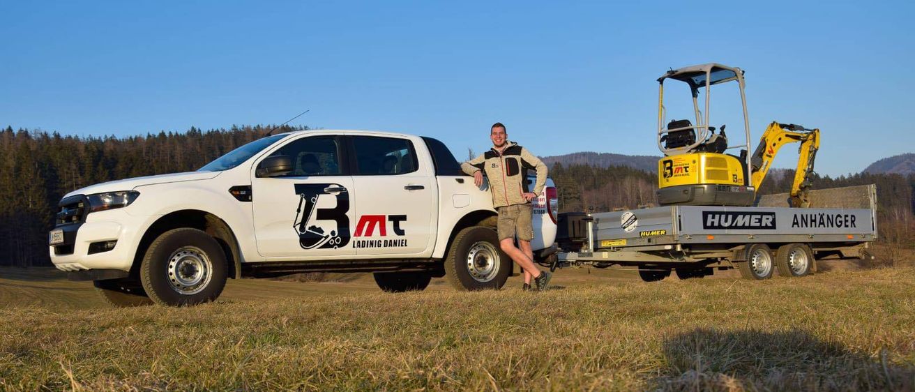 Ein Mann steht neben einem weißen Pickup-Truck mit einem Logo auf der Seite auf einem offenen Feld mit einem Baufahrzeug hinter ihm.