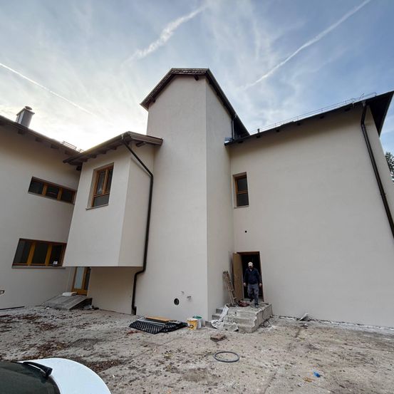 Ein modernes, weißes Gebäude im Bau mit einer Person an der Tür. Eine Leiter, Eimer und andere Materialien liegen auf dem Boden. Der Himmel ist klar mit ein paar Wolken.