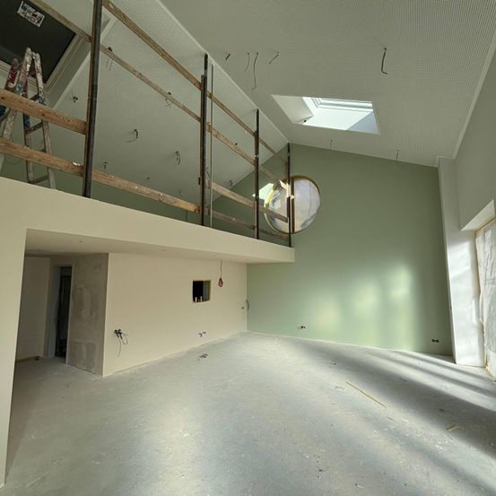 Ein unfertiger Raum mit grünen Wänden und einem Balkon mit Holzgeländer. Eine Leiter lehnt an der Wand. Der Raum hat eine weiße Decke mit einem Dachfenster und einem runden Fenster an der Wand.