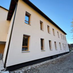 Ein modernes Gebäude mit beigefarbener Außenseite und schwarzem Rand. Das Gebäude hat viele Fenster mit Holzrahmen. Es gibt einige Steine auf dem Boden.