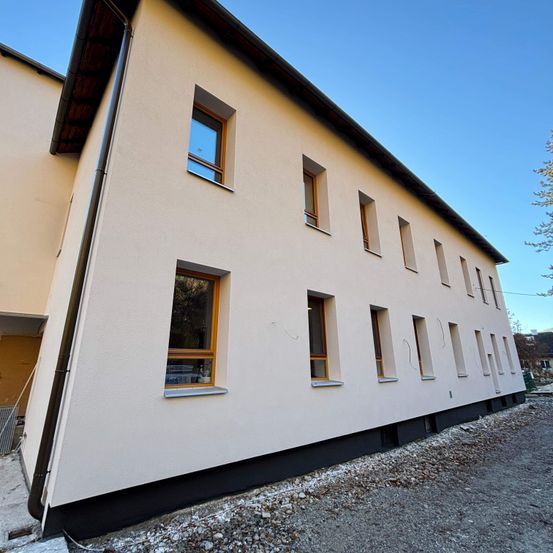Ein modernes Gebäude mit beigefarbener Außenseite und schwarzem Rand. Das Gebäude hat viele Fenster mit Holzrahmen. Es gibt einige Steine auf dem Boden.