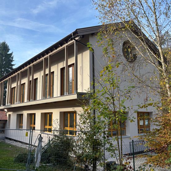 Ein modernes Gebäude mit beigefarbenen Wänden und gelben Fensterrahmen, es hat ein rundes Fenster an der Fassade und mehrere rechteckige Fenster auf jeder Etage. Das Gebäude ist von Bäumen und einem Zaun umgeben.