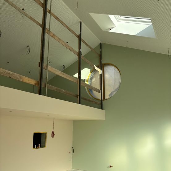 Ein unfertiger Innenraum mit Holzgerüst und weißer Decke. Ein großes transparentes Objekt hängt von der Decke. Ein kleines Fenster ist an der Wand.