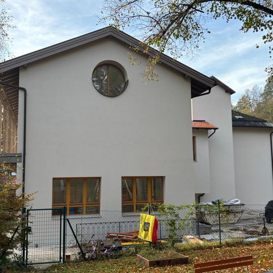Ein weißes Haus im Bau mit einem runden Fenster, einer gelb-roten Flagge und einem Zaun davor. In der Nähe befindet sich eine Holzbank und verstreute Gegenstände.