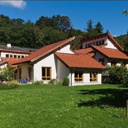 Ein großes weißes Haus mit rotem Dach steht in einem üppigen grünen Feld. Bäume umgeben das Grundstück, und ein blauer Stuhl ist draußen platziert.