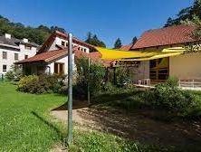 Ein Haus mit rotem Dach und gelbem Sonnensegel steht in einem üppigen grünen Garten mit Bäumen und Büschen unter einem klaren blauen Himmel.