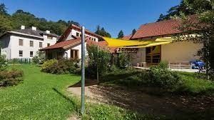 Ein Haus mit rotem Dach und gelbem Sonnensegel steht in einem üppigen grünen Garten mit Bäumen und Büschen unter einem klaren blauen Himmel.