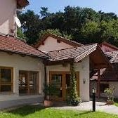 Ein weißes Gebäude mit rotem Dach und mehreren Fenstern steht vor einer Rasenfläche, mit Pflanzen und Bäumen im Hintergrund.