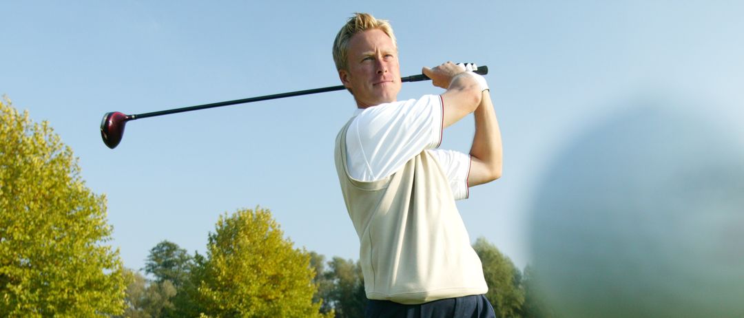 Ein Mann schwingt an einem sonnigen Tag einen Golfschläger. Er trägt ein weißes Shirt, schwarze Hose und Schuhe. Bäume und Gras umgeben ihn.