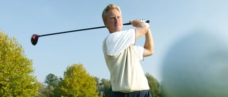 Ein Mann schwingt an einem sonnigen Tag einen Golfschläger. Er trägt ein weißes Shirt, schwarze Hose und Schuhe. Bäume und Gras umgeben ihn.