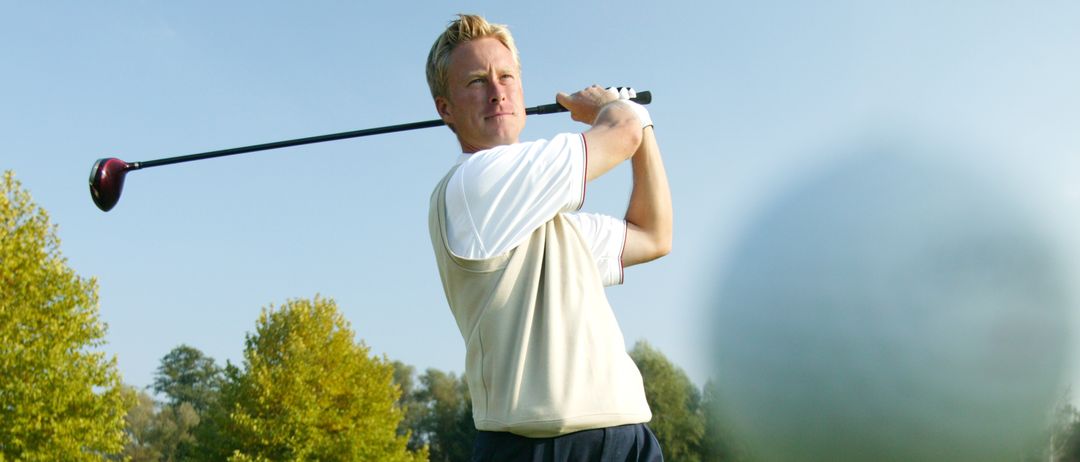 Ein Mann auf einem Golfplatz schwingt einen Schläger. Bäume und Gras umgeben den Golfer.