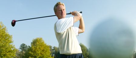 Ein Mann auf einem Golfplatz schwingt einen Schläger. Bäume und Gras umgeben den Golfer.