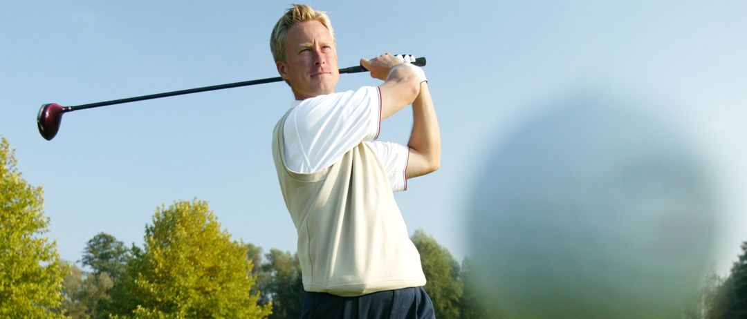 Ein Mann in einem weißen T-Shirt und schwarzen Hosen schwingt einen Golfschläger an einem sonnigen Tag auf einem Golfplatz.