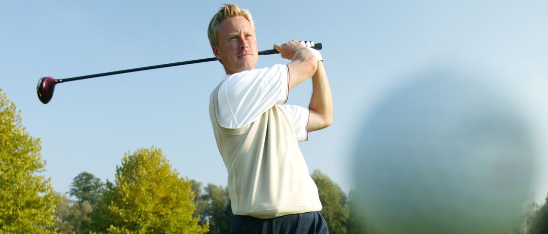 Ein Mann schwingt an einem sonnigen Tag einen Golfschläger und trifft den Ball weit. Grüner Rasen und Bäume im Hintergrund.