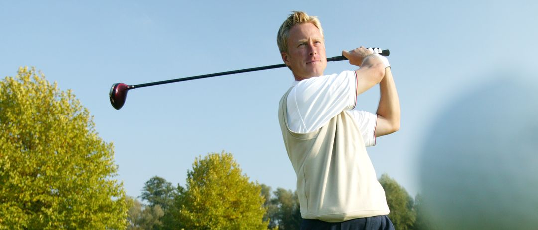 Ein Mann schwingt einen Golfschläger auf einem Golfplatz mit grünem Gras und Bäumen im Hintergrund.