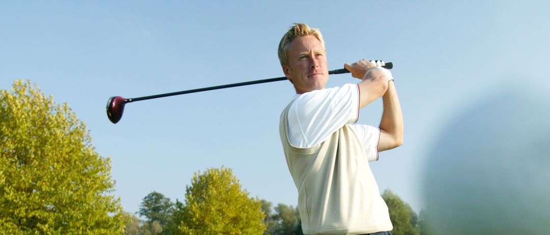 Ein Mann in weißem Shirt und dunkler Hose schwingt einen Golfschläger auf einem Golfplatz mit grünem Gras und Bäumen im Hintergrund.