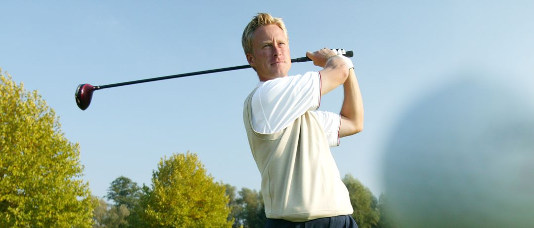 Ein Mann schlägt einen Golfball an einem sonnigen Tag auf einem Golfplatz. Er trägt ein weißes Shirt und dunkle Hosen.