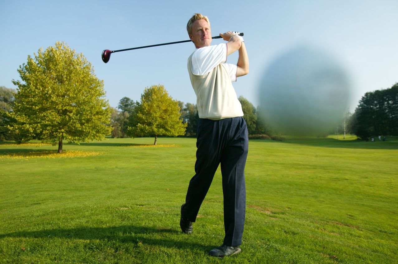 Ein Mann in einem weißen Shirt und dunkler Hose schwingt einen Golfschläger auf einem Golfplatz.