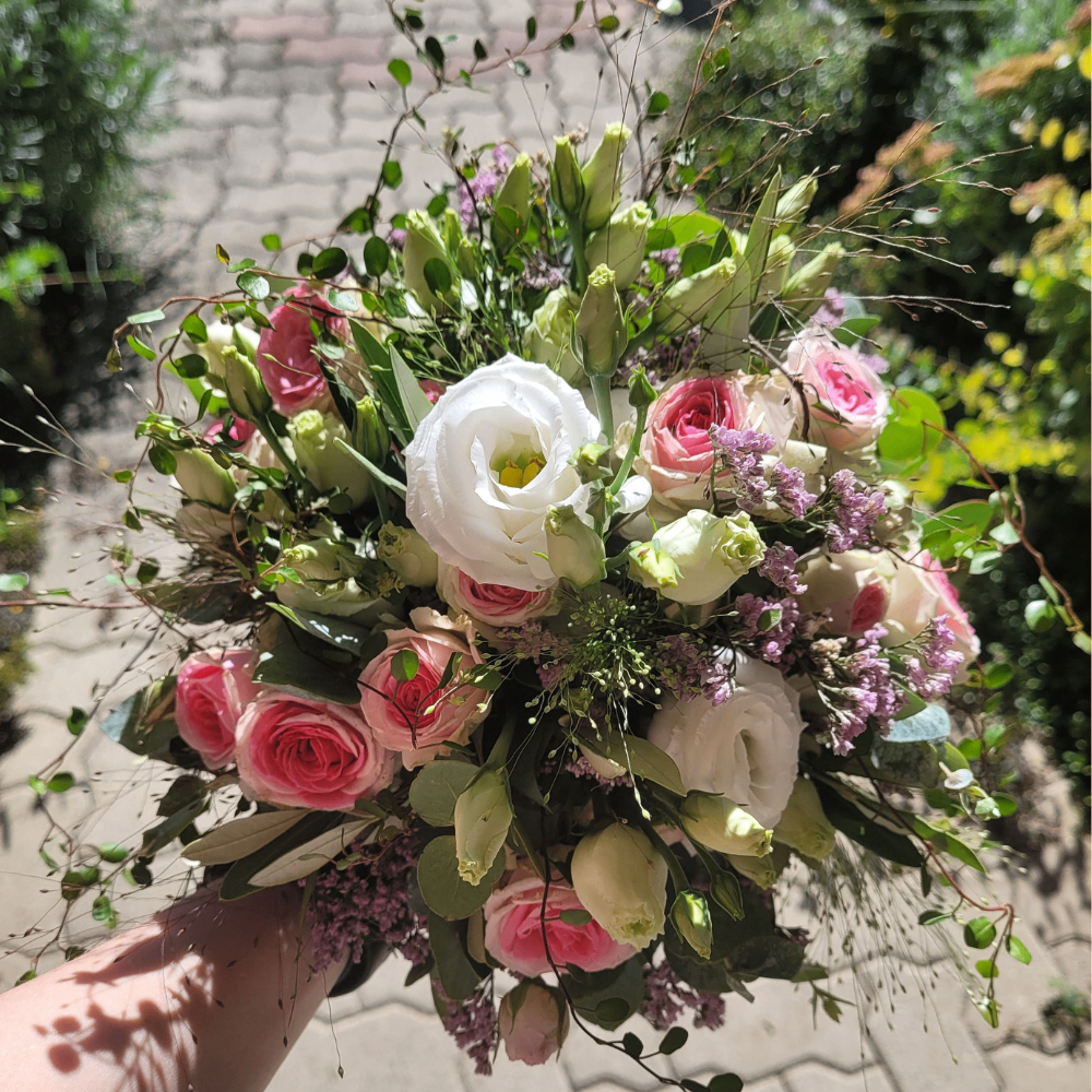 Eine Hand hält einen großen Strauß aus rosa und weißen Rosen mit grünen Blättern und lila Blumen. Der Strauß ist von grünem Laub umgeben.