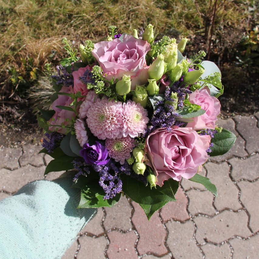 Eine Hand hält einen Blumenstrauß mit rosa Rosen, lila Blumen und grünen Blättern im Freien auf einem Ziegelweg.