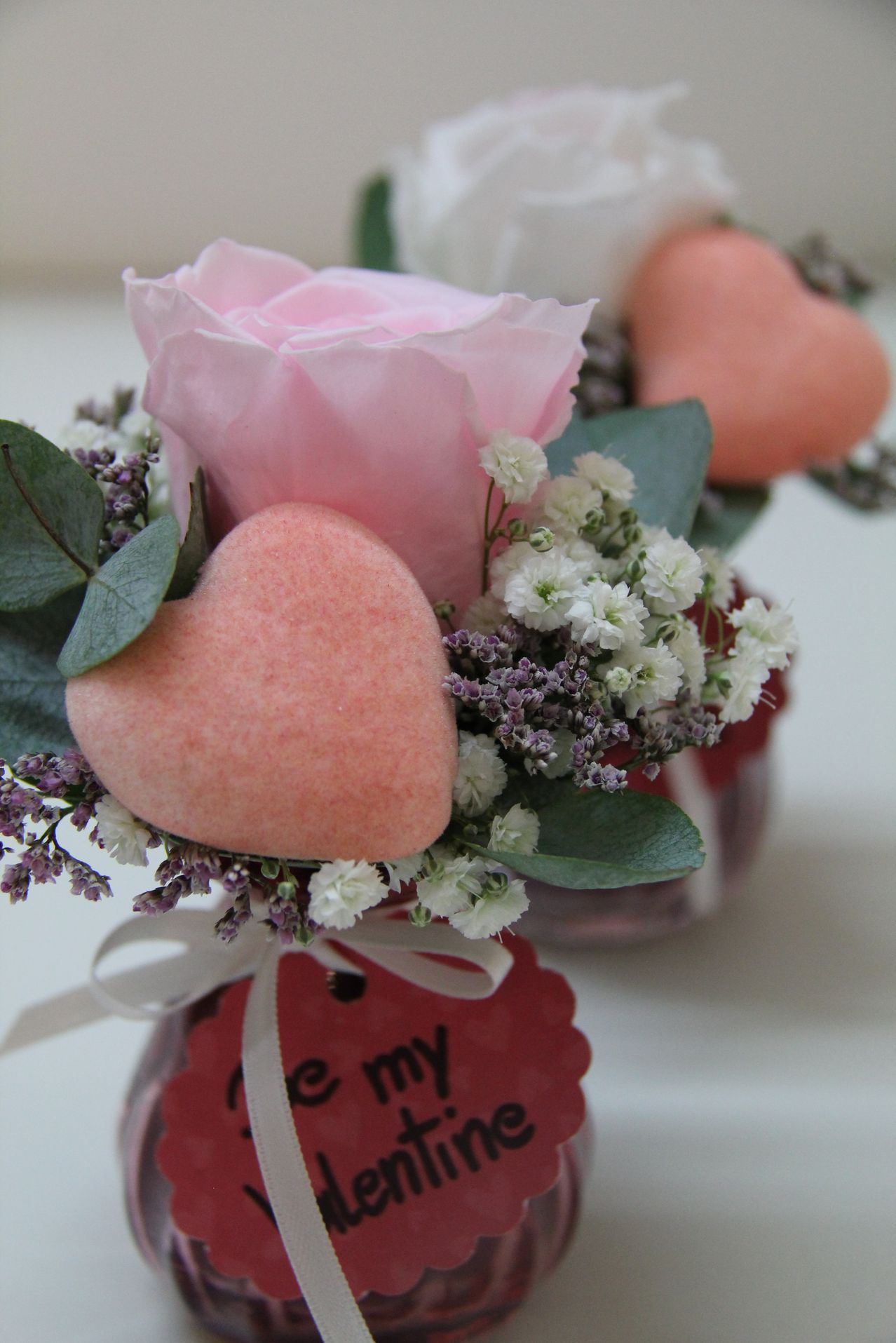 Eine rosa Rosenblume mit kleinen weißen Blüten und grünen Blättern steht in einem Vase mit einem herzförmigen Schwamm und einem Schild, das 'Sei mein Valentin' sagt.