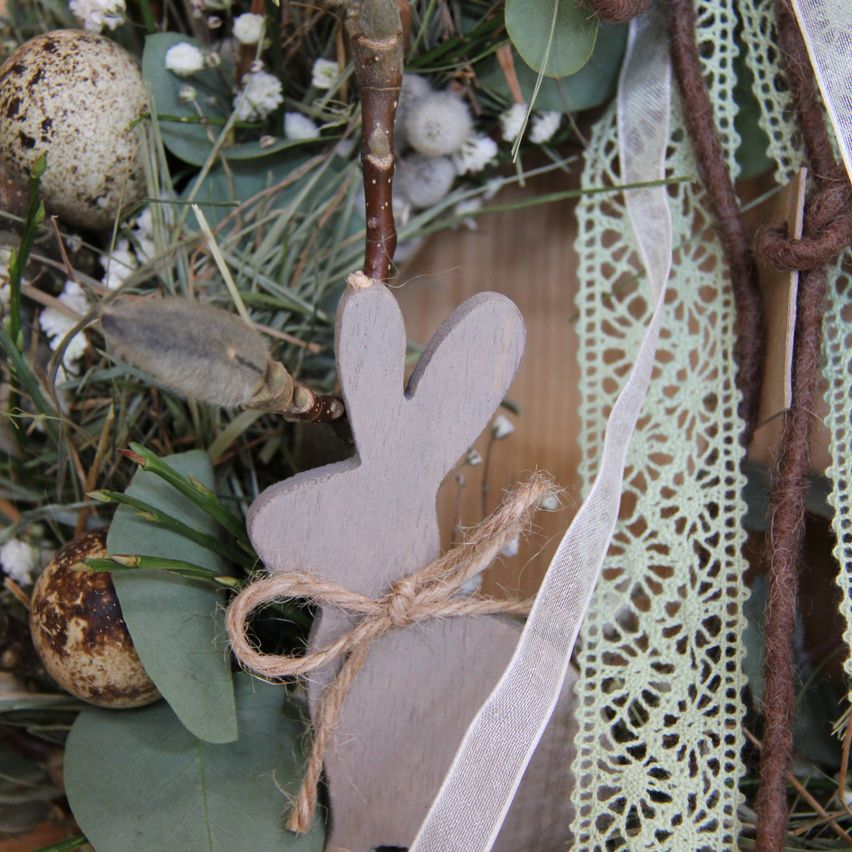 Ein hölzerner Hase mit einer braunen Schleife ist zwischen grünen Blättern, Zweigen und brauner Spitze platziert. Der Hintergrund besteht aus gefleckten Eiern und kleinen weißen Blüten.
