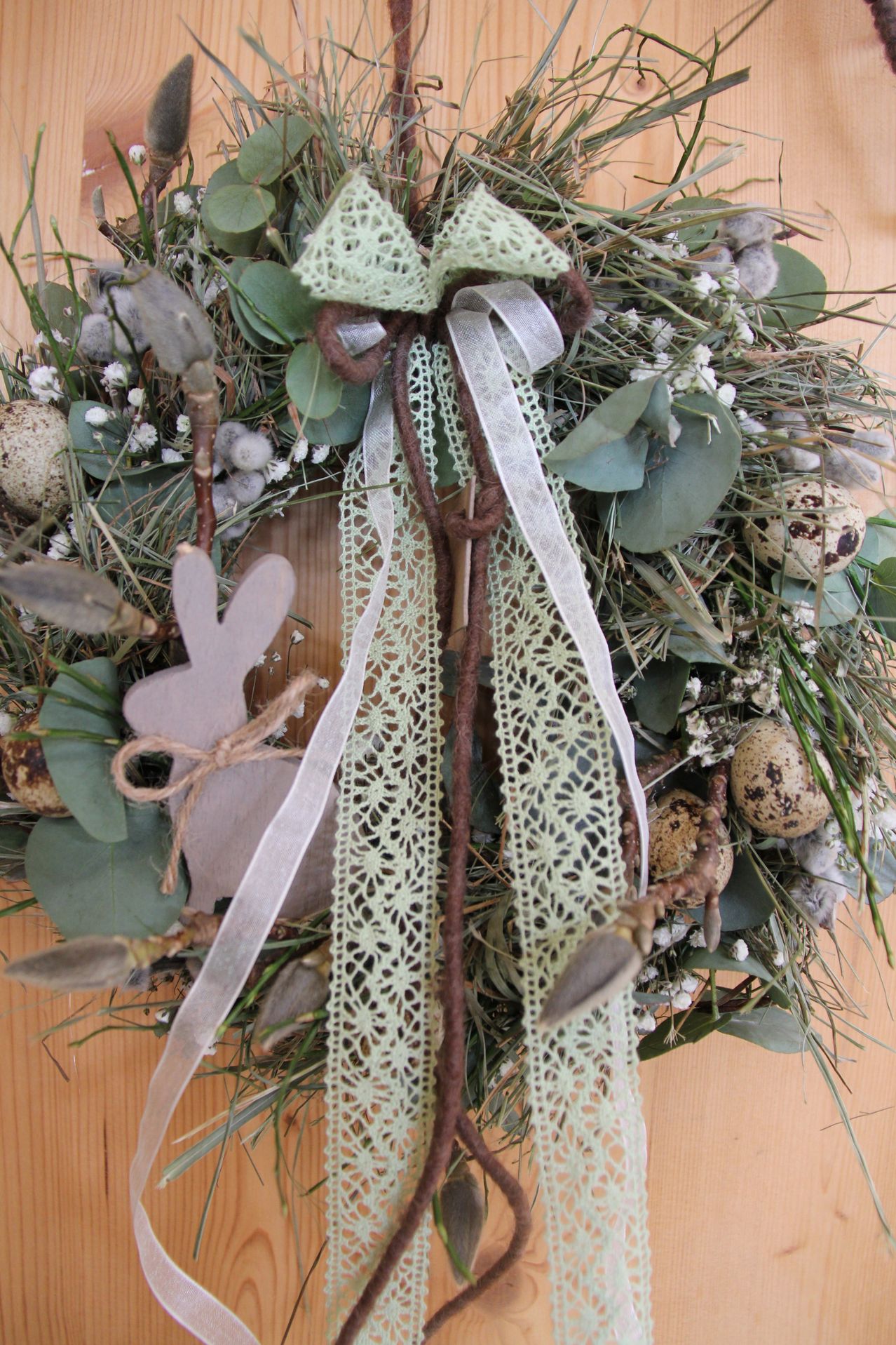 Ein Kranz mit einem weißen Band und einem dekorativen Hasen. Er hat grüne Blätter, kleine Eier und ein Spitzendesign. Der Kranz ist für den Frühling gemacht und steht auf einer Holzoberfläche.