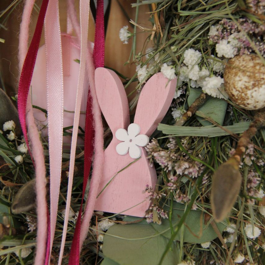Ein rosafarbener Holzhase mit einer weißen Blume auf dem Kopf befindet sich in einer Girlande aus Gras und kleinen Blumen.