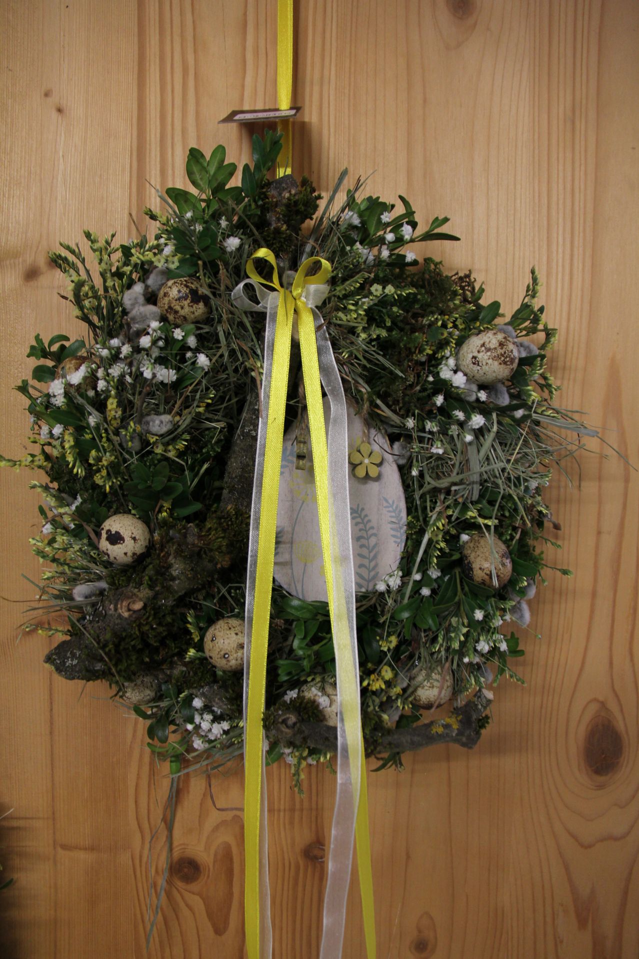 Ein Kranz aus Grün und Eiern hängt an einer Holzwand. Er hat ein gelbes Band und beinhaltet weiße Blumen und ein Blattdesign.
