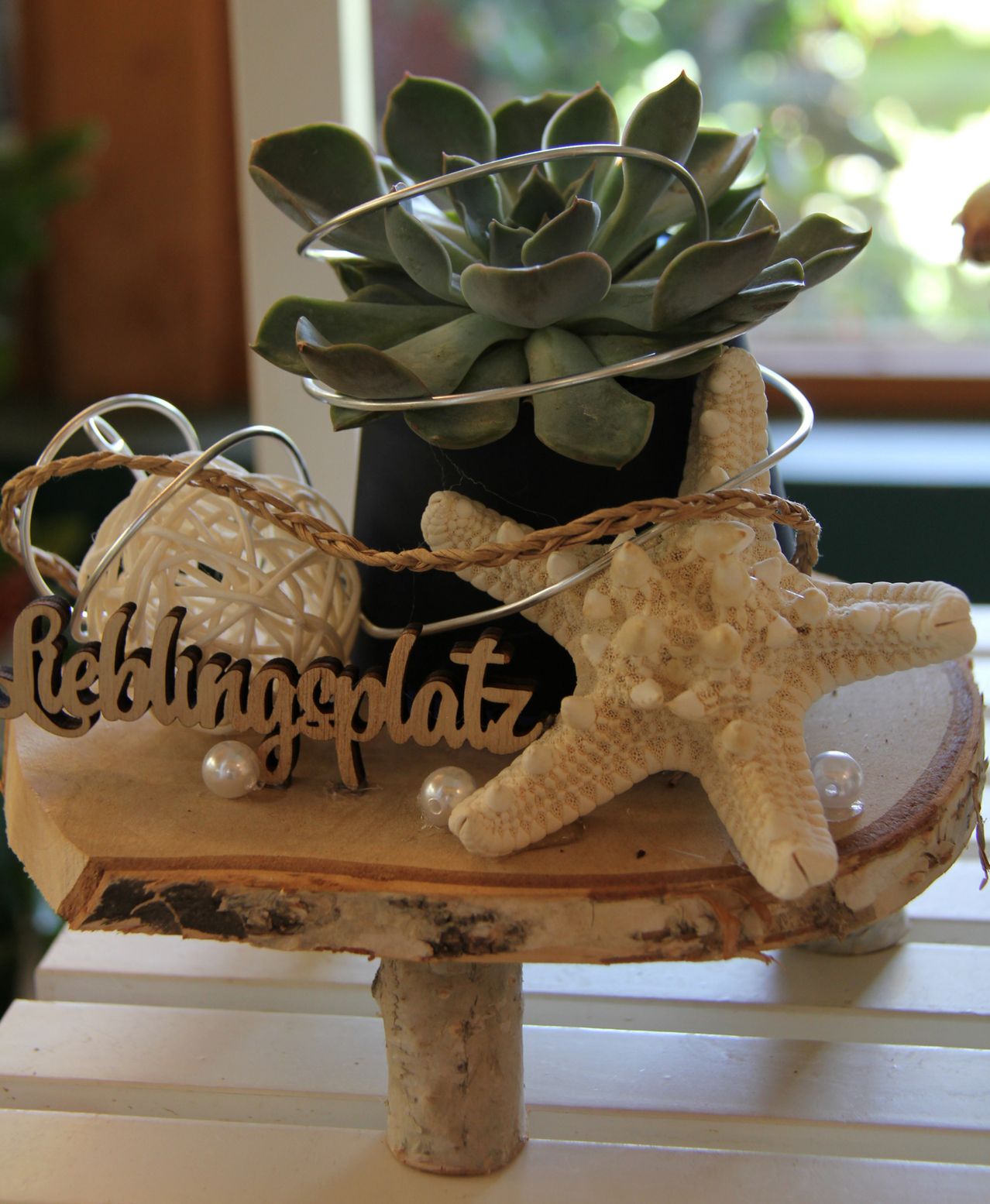 Ein Holztisch hält einen Seestern, eine dekorative Pflanze und ein handgefertigtes Schild mit der Aufschrift 'Reblingtonplatz', mit einer Perle und einem Weidenkorb.