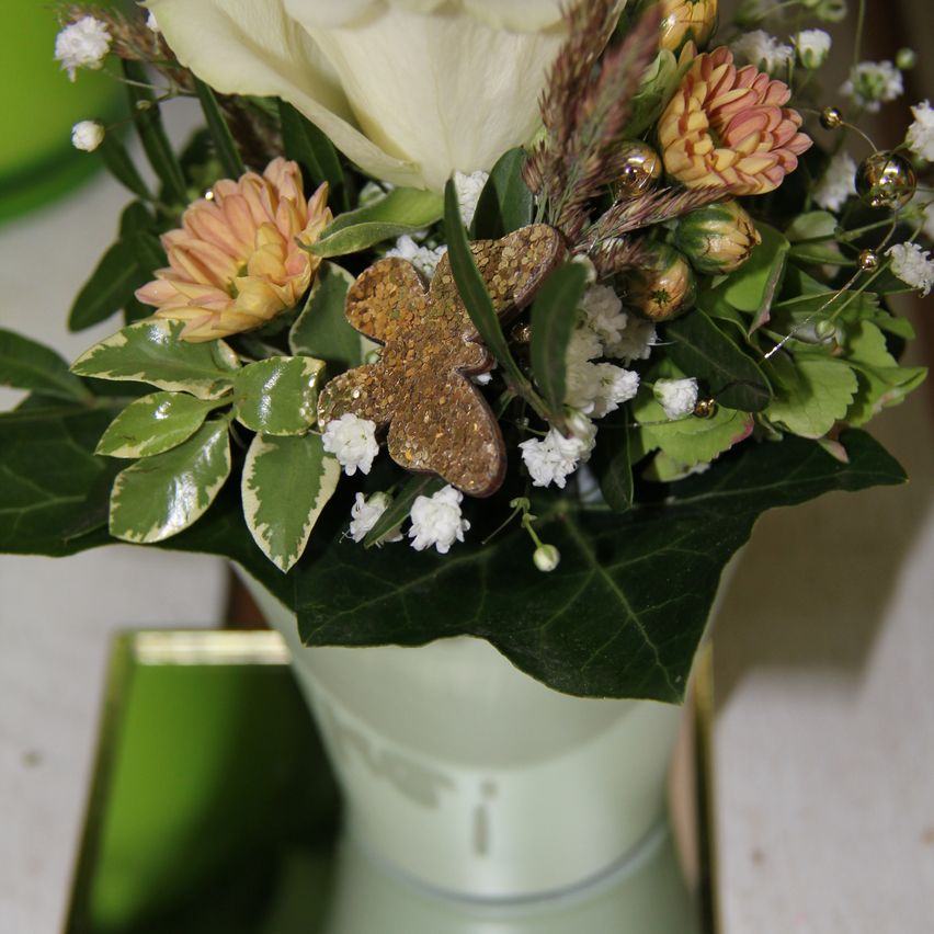 A green vase holds a floral arrangement with a white rose, yellow and white daisies, and various green leaves. The arrangement includes decorative elements like shiny gold pieces.