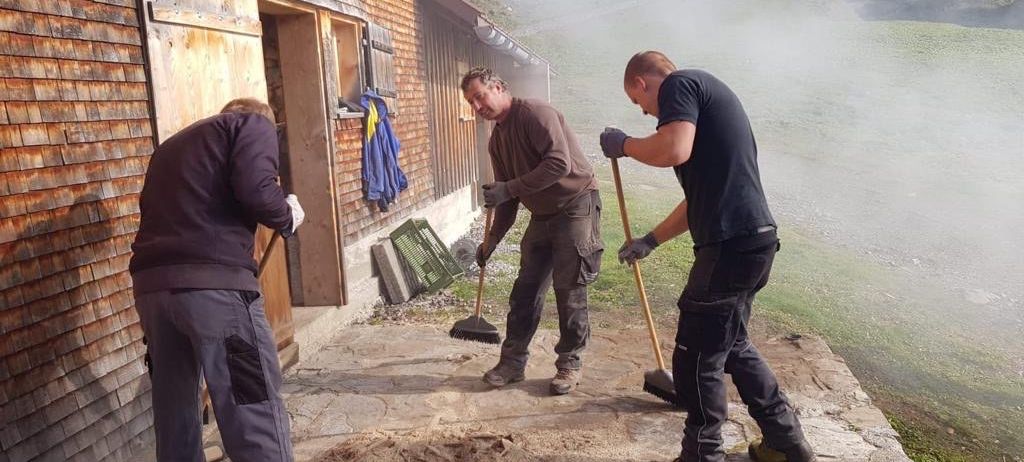 Drei Männer in Arbeitskleidung reinigen einen Steinweg vor einem Holzgebäude. Sie halten Besen in der Hand und konzentrieren sich auf ihre Aufgabe.