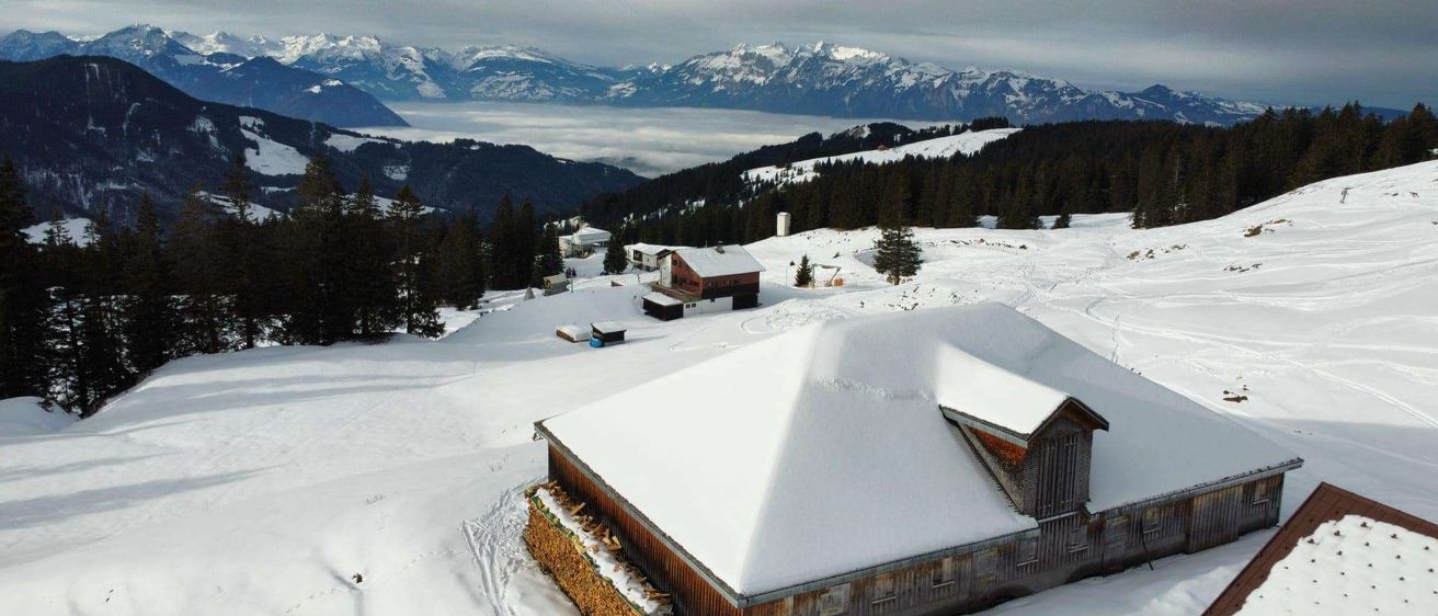 Eine verschneite Landschaft mit einem Holzschuppen und einer Bergkette im Hintergrund. Bäume bedecken die unteren Hänge.