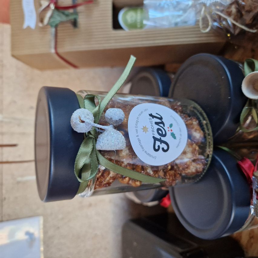 Ein Glas mit schwarzem Deckel und grüner Schleife enthält festliche Kekse. Dahinter befinden sich weitere Gläser und eine Holzkiste mit Weihnachtsdekoration.